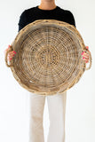 A person holding a large round woven wicker basket with side handles against a plain white background.