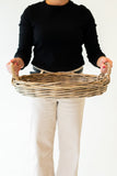 A person wearing a black long-sleeve top and cream pants holds a round woven wicker basket with two handles against a plain white background.
