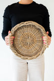 A person wearing a black long-sleeve top and white pants holds a round woven wicker basket tray with side handles against a plain white background.
