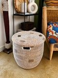 A round woven storage basket with a lid, made from natural beige and brown fibers, placed on a beige carpet in a styled room next to a wicker chair.
