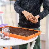 Person holding seeds over a tray of soil with a jar on a table.