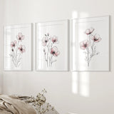 Three floral wall art prints on a white wall with a neutral-colored blanket and plant in the foreground.