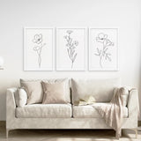 Beige sofa with decorative pillows and a blanket, featuring three framed floral line art prints on the wall.