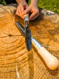 A close-up of a hand holding a Sharp Shooter Trowel with a dark metal blade and light wood handle, resting on a large tree stump outdoors. Another similar tool lies on the stump below.