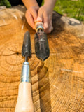 A close-up of a person's hands holding a dark metal Sharp Shooter Trowel over a large cut tree stump outdoors, with another hand tool resting on the wood.