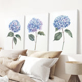 Three panels of blue hydrangea flowers on a wall above a bed with pillows and a lamp.