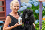 Woman holding a bottle of TUBEROSE GOATS MILK LOTION next to a black dog with flowers in the background