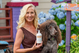 Woman holding a dog and a bottle of 'Good' product in front of flowers and a colorful background.