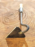 A close-up of a V-Hoe (Warren Hoe, Aggressive Hoe) Hand Forged Garden Tool hand tool with a triangular metal blade and a curved metal handle ending in a light wood grip, resting on a textured wooden surface.