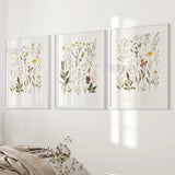 Three framed botanical prints on a wall with a bed and blanket in the foreground.