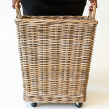 A natural woven wicker laundry hamper with rope handles and small black caster wheels, shown from the front with a person's hands holding the handles at the top.