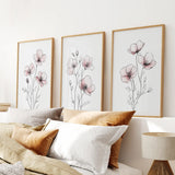 Three floral prints on a wall above a bed with pillows and a lamp.