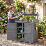 A woman in an apron and gloves tends to a potted plant beside a large gray outdoor garden storage cabinet with shelves and hanging tools, set in a stone patio garden courtyard.