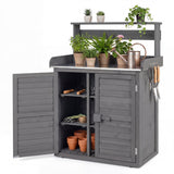 A dark gray wooden outdoor garden storage cabinet with a built-in work surface, top shelf, open lower cabinet, and hanging gardening tools. The cabinet is shown with one door open, revealing shelves with pots and tools, and the top surface decorated with potted plants, a watering can, and a rake, on a white background.