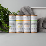 Four J.J. Naturals deodorant cans on a shelf with towels and a plant in the background.