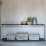 A black metal console shelf with open lower shelves, styled with a potted plant, a wooden bowl, a gray ceramic jar, and three wicker storage baskets against a textured beige wall.