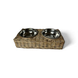 A rectangular woven wicker pet feeder stand holding two round stainless steel bowls, photographed on a clean white background.