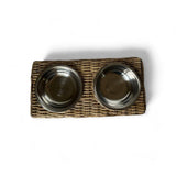 A rectangular woven wicker pet feeder tray holding two round stainless steel bowls, photographed from above on a white background.