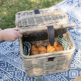 A natural wicker picnic basket with a hinged lid is shown outdoors on a blue-and-white patterned blanket, with a hand lifting the lid. Croissants are visible inside, along with small green-and-white check cloth accents.