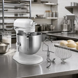 A white KitchenAid stand mixer with a stainless steel bowl sits on a stainless steel kitchen counter, with two attachments placed beside it. The background shows a professional kitchen environment with racks, bowls, and another mixer.