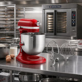 A red KitchenAid stand mixer with a stainless steel bowl sits on a commercial kitchen counter, with a metal hook and wire whisk attachment beside it. In the background is a large stainless steel commercial oven, with eggs, milk, and a bowl of flour on the counter.