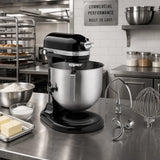 A black KitchenAid stand mixer with a stainless steel bowl sits on a stainless steel kitchen counter, surrounded by baking ingredients and mixing attachments in a commercial kitchen setting.