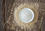 White bowl of rice flour surrounded by raw rice on a rustic wooden surface