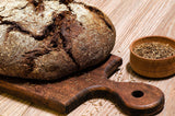 Rustic loaf of bread on a wooden cutting board with a small bowl of seeds on a wooden surface.
