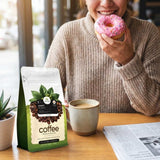 Person holding a pink donut with a coffee bag and cup on a table