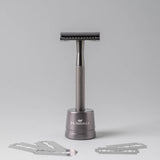 A dark gray metal safety razor with a textured grip and a matching cylindrical stand, shown on a light gray background with four razor blades arranged in front. The stand is branded with the JHU NATURALS logo.