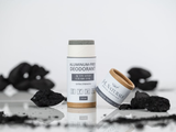Two deodorant sticks from 14 Naturals are displayed on a reflective white surface with loose black charcoal pieces scattered around them. One stick is upright and shows a white label with gray and dark gray text, while the other lies on its side showing a kraft paper tube with a white label.
