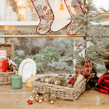 A styled holiday tabletop scene with woven baskets, hanging Christmas stockings, a small artificial tree, red plaid ribbon, pinecones, ornaments, and a red mug on a wooden surface in front of a window.