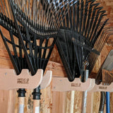 Set of black garden rakes hanging on a wooden wall with 'Hangs This Up' tool holder.