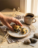 Person holding a blueberry muffin with a cup of coffee on a wooden table.