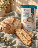 A bag of af Jules gluten free sourdough and baking flour stands on a textured blue-gray cloth beside a round rustic loaf of bread and two sliced pieces, with warm natural light and a softly blurred background.