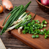 Fresh green onions, including whole unchopped stalks and finely chopped pieces, arranged on a wooden cutting board with whole and sliced red onions in the background.