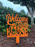 Orange 'Welcome to our Haunted House' sign on a stand with greenery in the background