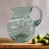 Glass pitcher with swirling design on a wooden surface with green apples in the background