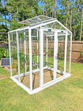 White framed glass greenhouse in a backyard with trees and a solar panel in the background. The Sago Greenhouse 5' x 8'