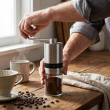 Person using a manual coffee grinder on a wooden table with coffee beans and a cup.