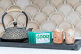 Tea kettle, candles, and soap on a tray with a decorative wall in the background