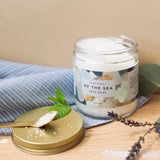 Jar of bath salts labeled 'Of the Sea' on a wooden surface with a spoon and mint leaves.