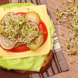 Sandwich with lettuce, tomato, cheese, and pickles on a wooden surface with sprouts.