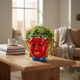 Decorative plant pot shaped like a red face on a wooden table with a blurred indoor background