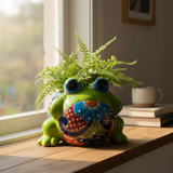 Colorful frog-shaped planter with a plant on a wooden surface near a window.