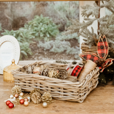 A natural woven wicker basket filled with pinecones and twine sits on a wooden surface in a rustic holiday setting, with red plaid ribbon, gold ornaments, and a blurred outdoor window background.