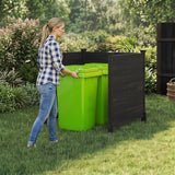 A woman in a blue and white plaid shirt and jeans stands next to a black plastic bin enclosure with two bright green bins in a grassy backyard garden setting.