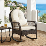 A dark brown wicker rocking chair with cream cushions sits on a woven rug on a wooden deck, next to a small black side table, with a scenic ocean and house view in the background.