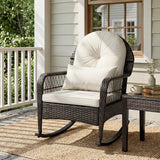A dark brown wicker rocking chair with cream cushions sits on a porch next to a matching side table. The chair is positioned on a woven jute rug in front of a house with light beige siding and a window.